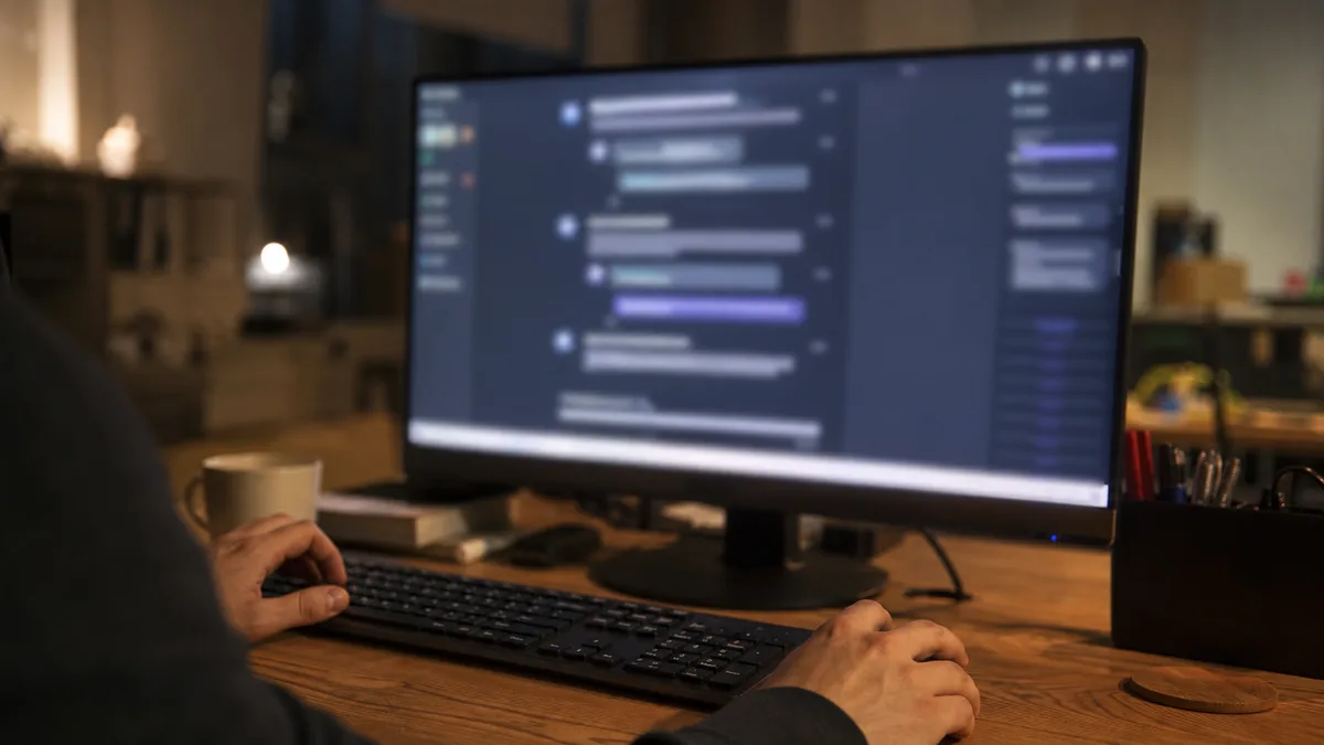 Pohled přes rameno zezadu na velký monitor s rozostřeným chatovacím rozhraním AI asistenta, osoba anonymní, večerní světlo kanceláře malé firmy