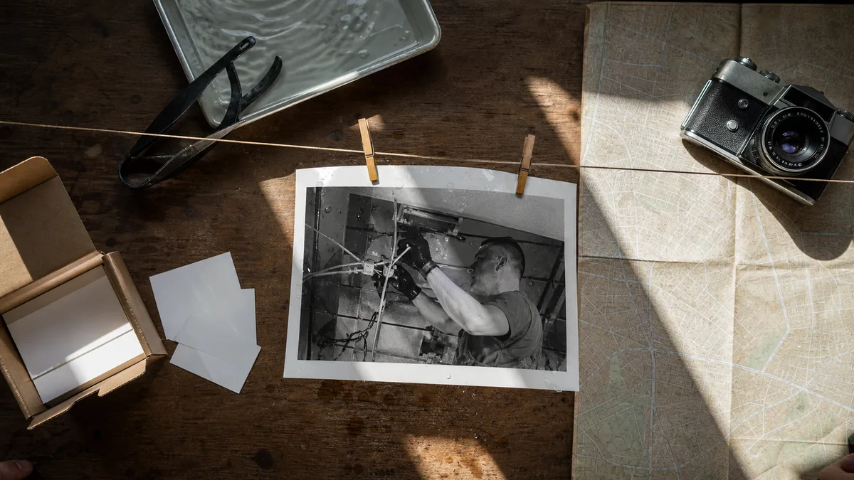 Temná komora: na šňůře schne vyvolaná černobílá fotografie elektrikáře při práci, vedle vývojová miska, kleště, mapa města a starý fotoaparát — obraz firmy se skládá ze stop, které po sobě nechává